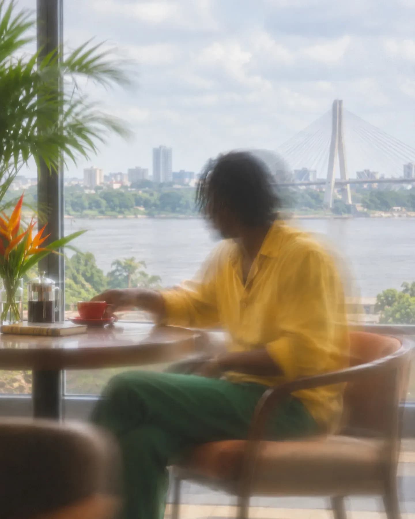 A man in a yellow shirt reaches for a red cup in a Brazzaville cafe overlooking the Congo River and Pont du 15 Aout 1960.
