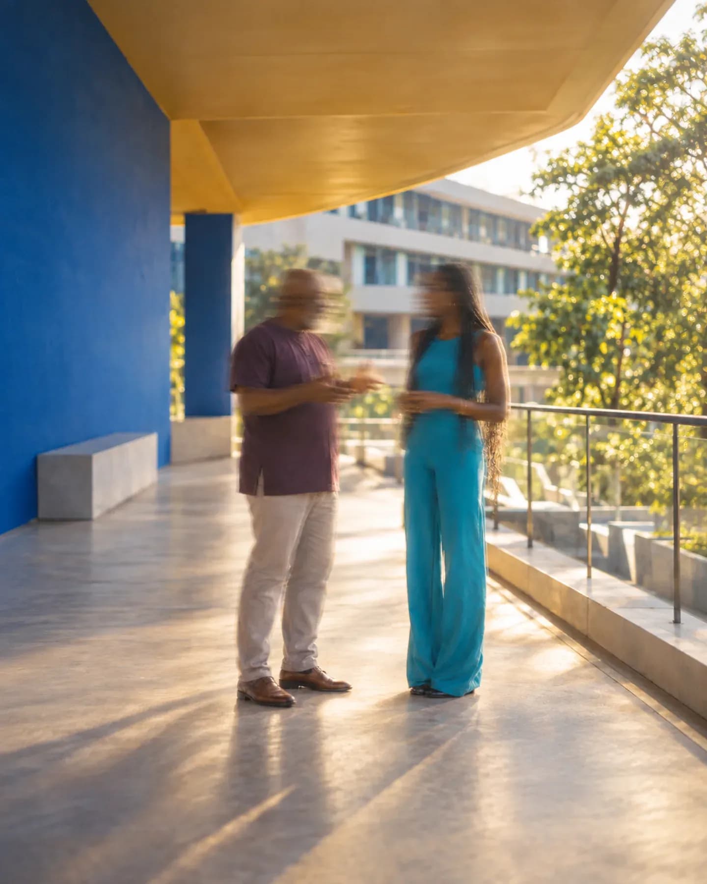 A man and a woman in a turquoise jumpsuit talk under a yellow canopy beside a blue wall, trees and buildings beyond.
