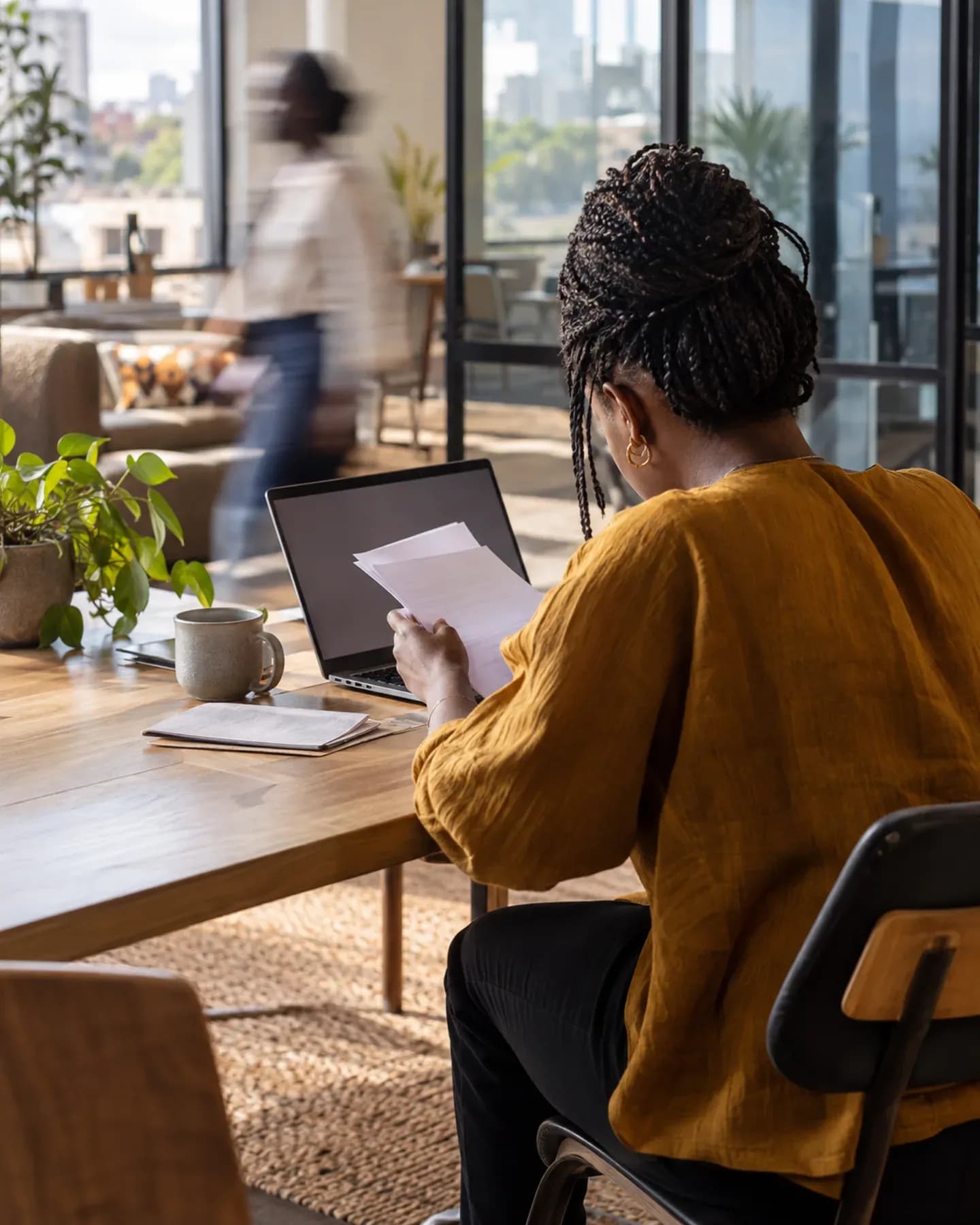 A researcher in an ochre blouse reviews papers beside a laptop in a bright contemporary office as a colleague passes behind her.