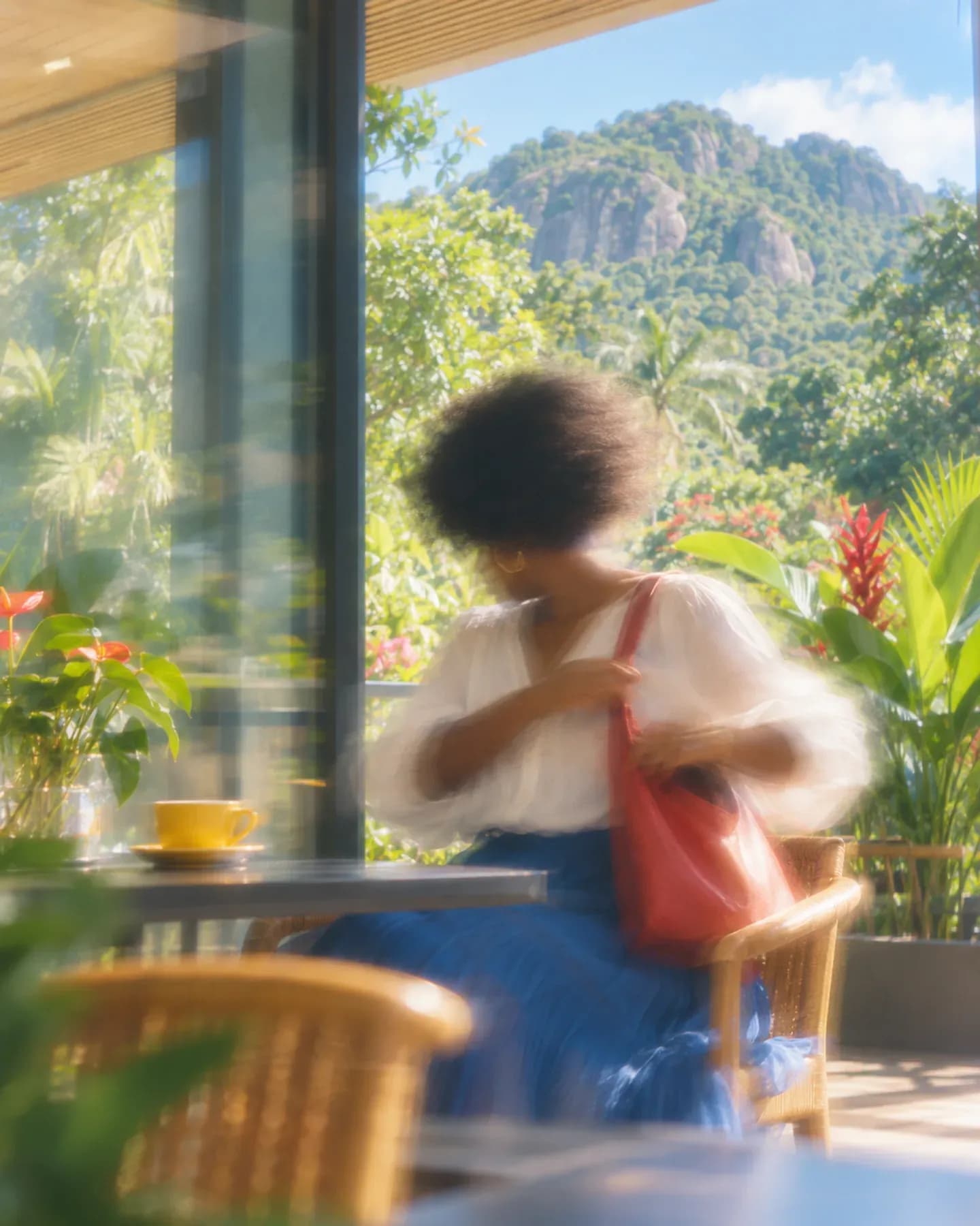 A woman in a white blouse adjusts a red shoulder bag in a Victoria cafe near Mont Fleuri, with green granite hills beyond.