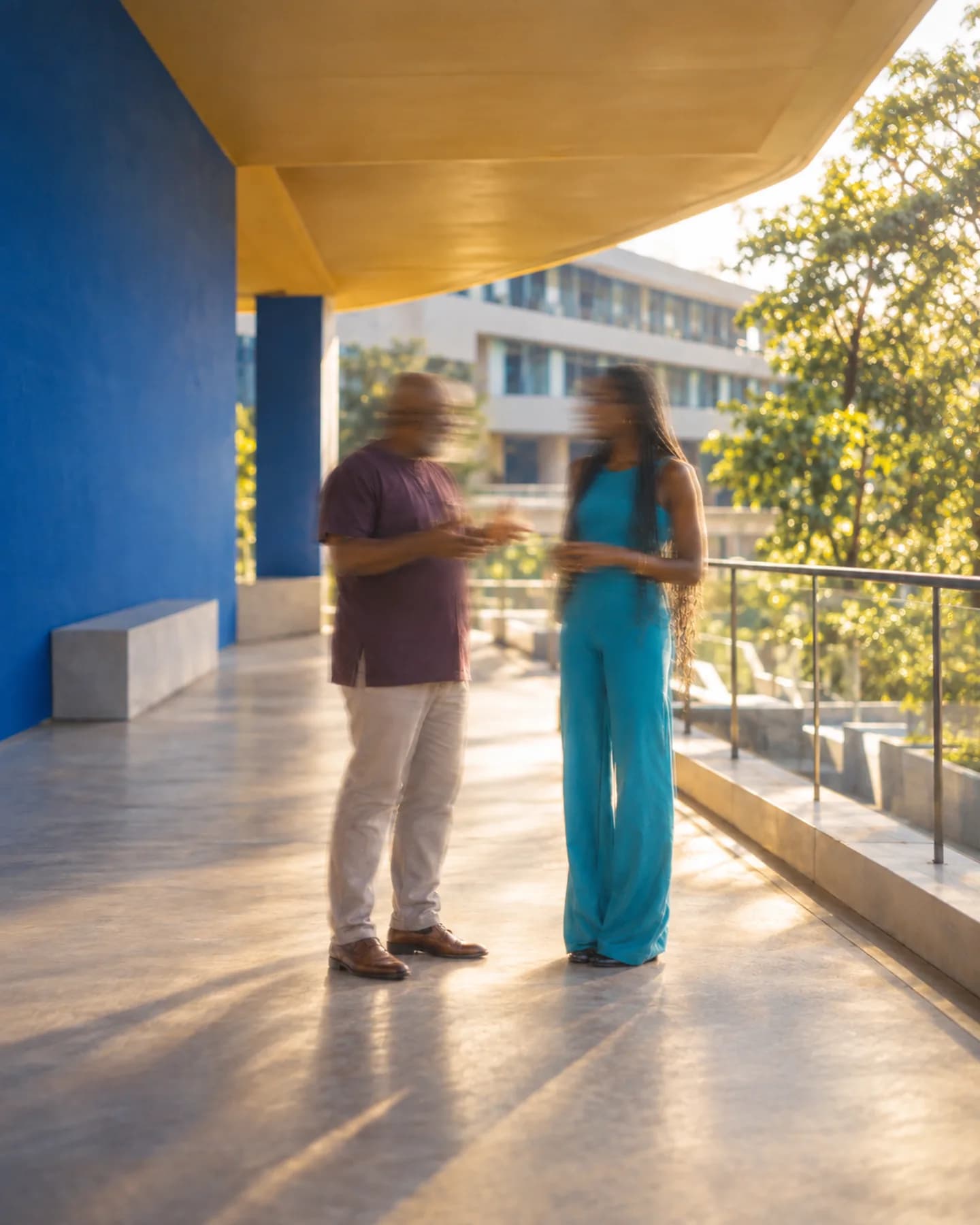 Un homme et une femme en combinaison turquoise discutent sous un auvent jaune près d'un mur bleu, avec des arbres et des immeubles au loin.